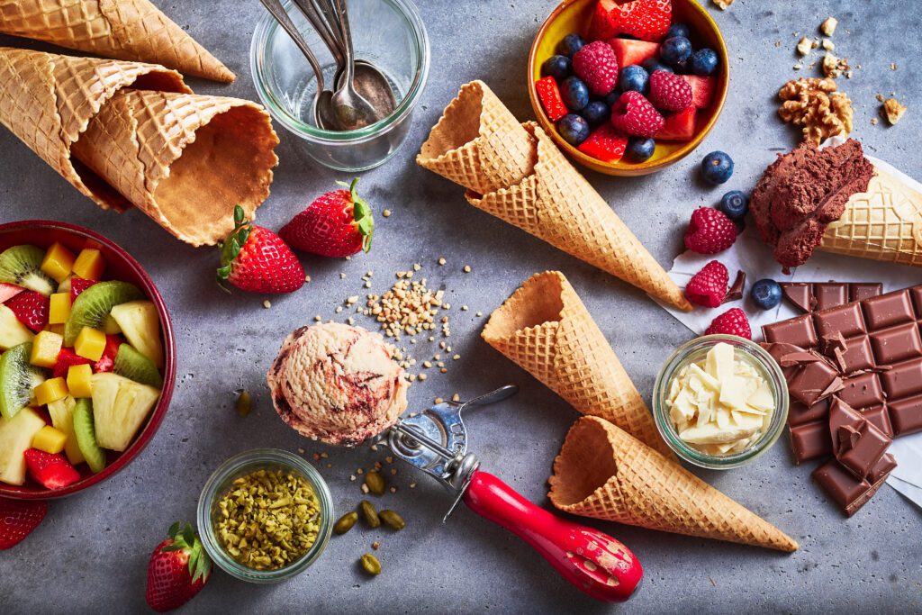 waffle cones and a variety of gelato ingredients, including pistachio, berries, tropical fruit, and chocolate laying on a table