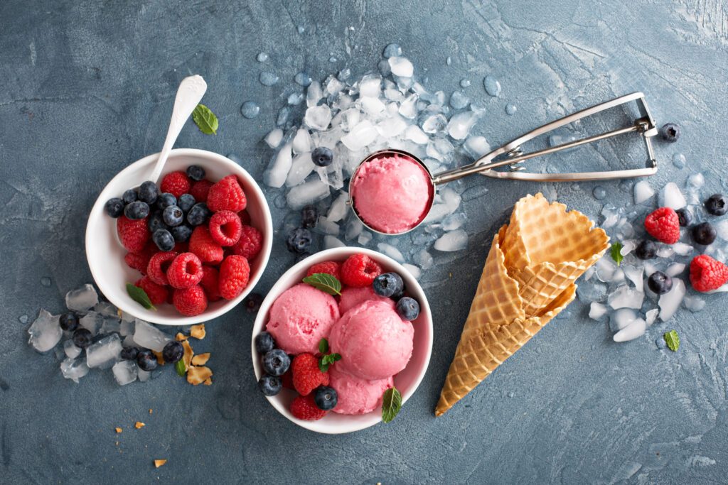 a bowl of mixed berries next to a bowl of bright pink gelato and waffle cones